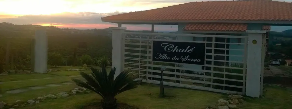 Portão de entrada da pousada ao pôr do sol, com a placa Chalé Alto da Serra e morros ao fundo