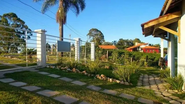 Caminho de pedras no jardim com cerca de metal, coqueiro e chalés ao fundo sob céu azul