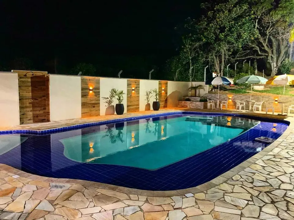 Piscina de azulejos azuis iluminada à noite, com deck de pedra e espreguiçadeiras ao fundo